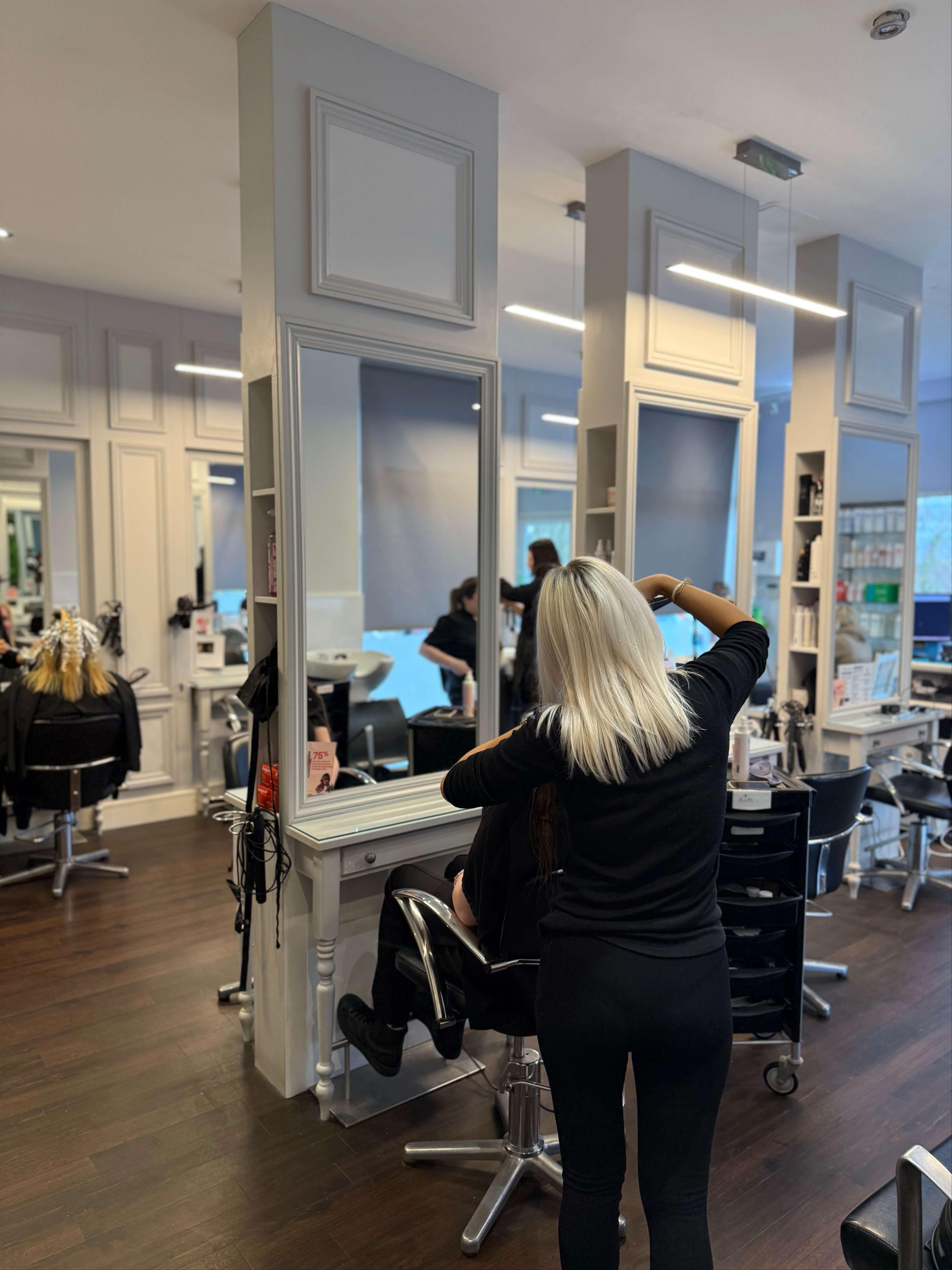 Hair stylist standing behind a seated client in a bright, modern hair salon, working on the client’s long blonde hair in front of a large framed mirror.